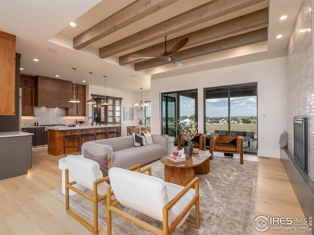 living room featuring a large fireplace, beamed ceiling, light wood-type flooring, and ceiling fan with notable chandelier