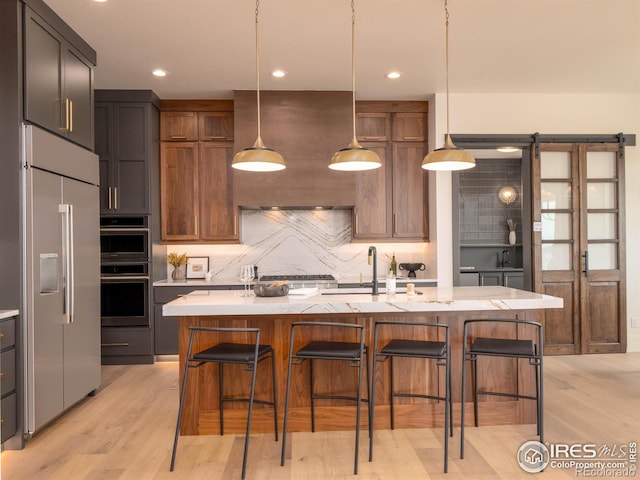 kitchen with a kitchen island with sink, a barn door, decorative light fixtures, light wood-type flooring, and appliances with stainless steel finishes