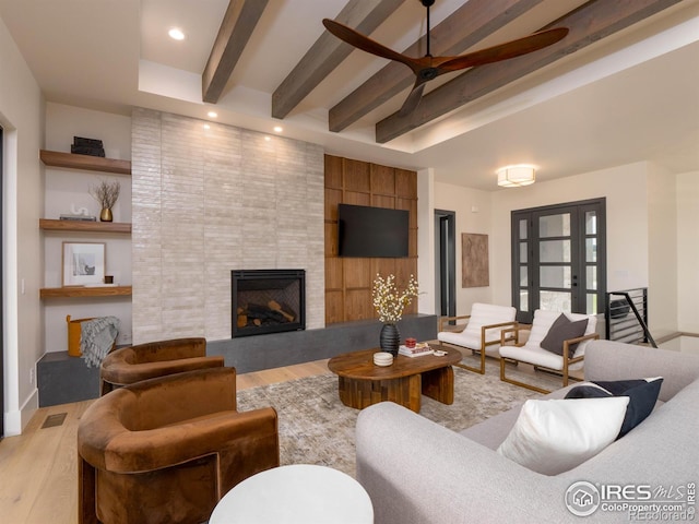 living room with a tiled fireplace, ceiling fan, light wood-type flooring, beamed ceiling, and built in features