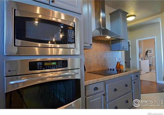 kitchen with appliances with stainless steel finishes, gray cabinetry, backsplash, wall chimney exhaust hood, and hardwood / wood-style floors