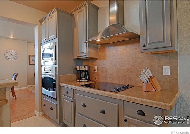 kitchen with light wood-type flooring, wall chimney range hood, gray cabinetry, stainless steel appliances, and backsplash