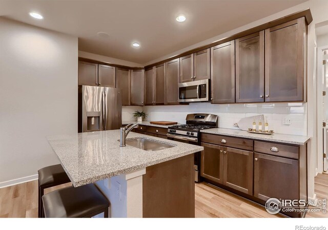 kitchen with light wood-type flooring, appliances with stainless steel finishes, light stone countertops, sink, and a kitchen island with sink