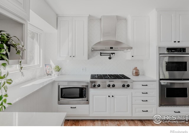 kitchen with white cabinetry, light hardwood / wood-style flooring, decorative backsplash, wall chimney exhaust hood, and appliances with stainless steel finishes