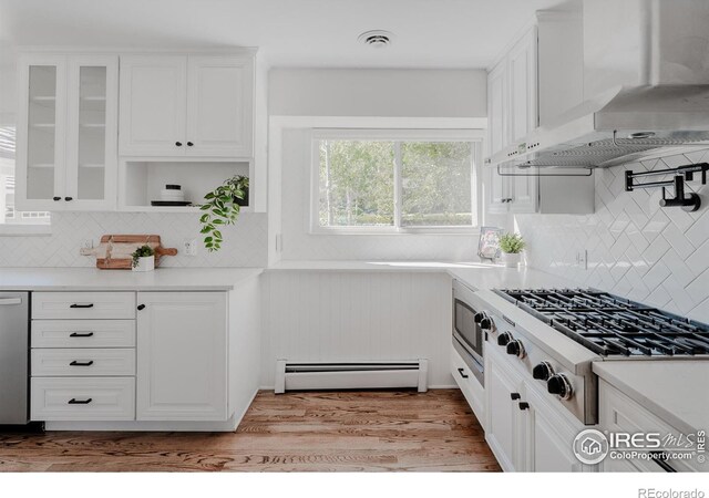 kitchen featuring light wood-type flooring, appliances with stainless steel finishes, white cabinetry, wall chimney range hood, and a baseboard radiator