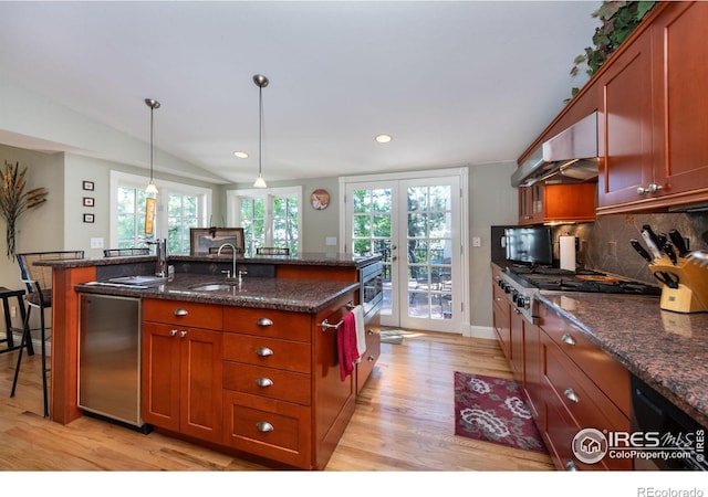 kitchen featuring range hood, an island with sink, sink, dark stone countertops, and hanging light fixtures