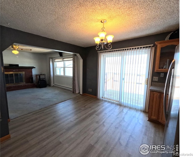 unfurnished dining area featuring ceiling fan with notable chandelier, a baseboard radiator, hardwood / wood-style flooring, a brick fireplace, and a textured ceiling