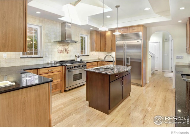 kitchen with high quality appliances, wall chimney exhaust hood, a tray ceiling, and a center island with sink