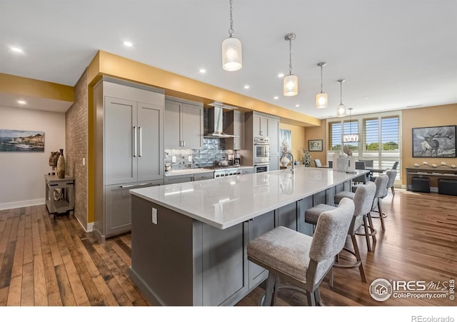 kitchen with gray cabinetry, decorative light fixtures, wall chimney exhaust hood, dark wood-type flooring, and a large island with sink