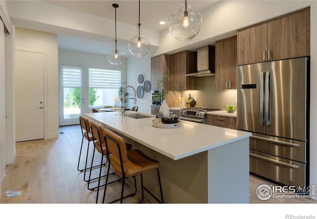 kitchen with premium appliances, wall chimney exhaust hood, an island with sink, and light hardwood / wood-style flooring