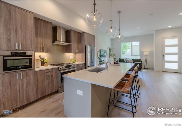 kitchen with light wood-type flooring, an island with sink, stainless steel appliances, and wall chimney range hood