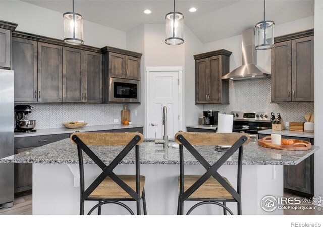 kitchen featuring a center island with sink, stainless steel appliances, wall chimney range hood, and a breakfast bar