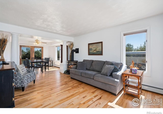 living area with light wood finished floors, a wood stove, a ceiling fan, decorative columns, and baseboard heating