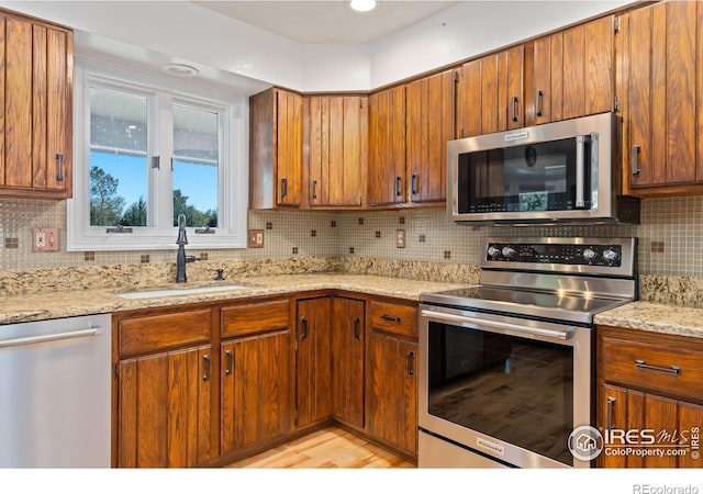 kitchen with stainless steel appliances, a sink, tasteful backsplash, brown cabinets, and light stone counters