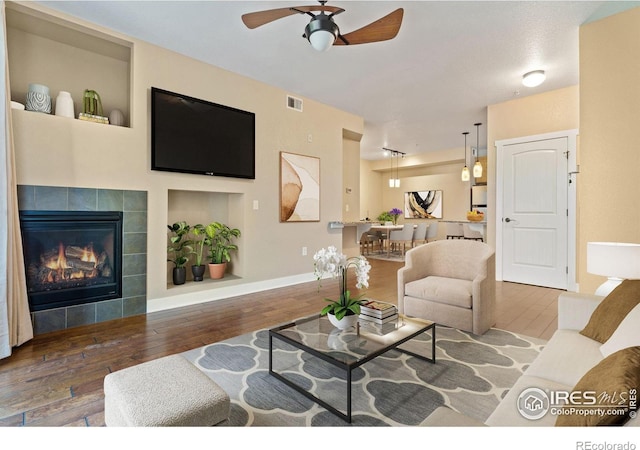 living room with hardwood / wood-style flooring, ceiling fan, built in shelves, and a tiled fireplace