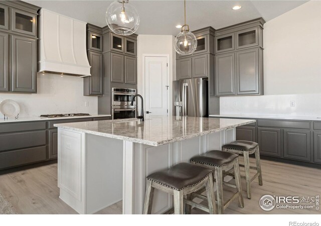 kitchen with custom exhaust hood, an island with sink, appliances with stainless steel finishes, light stone countertops, and light hardwood / wood-style floors