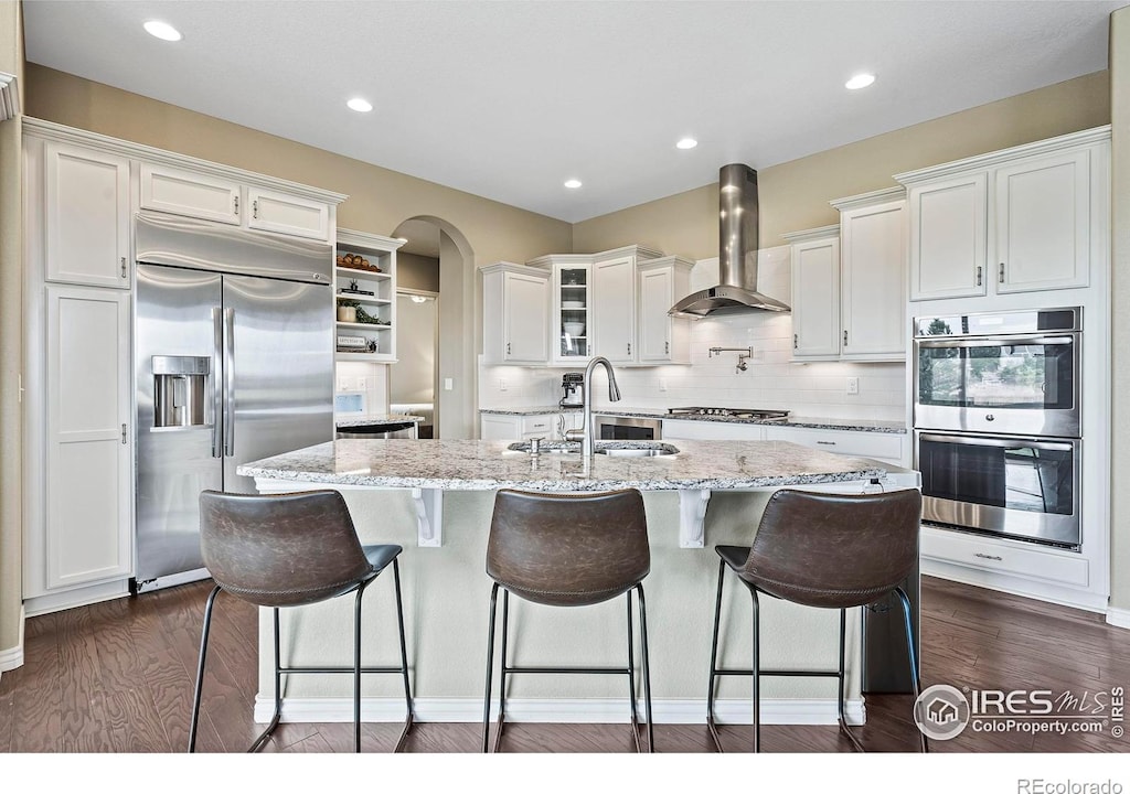 kitchen featuring white cabinets, a kitchen island with sink, dark wood-type flooring, wall chimney range hood, and stainless steel appliances