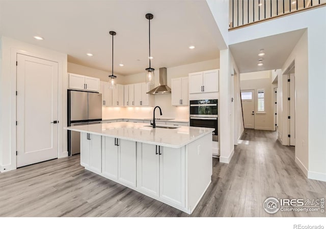 kitchen featuring white cabinets, stainless steel appliances, a kitchen island with sink, and wall chimney range hood