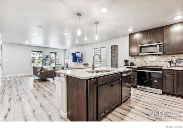 kitchen featuring an island with sink, hanging light fixtures, sink, stainless steel appliances, and light wood-type flooring