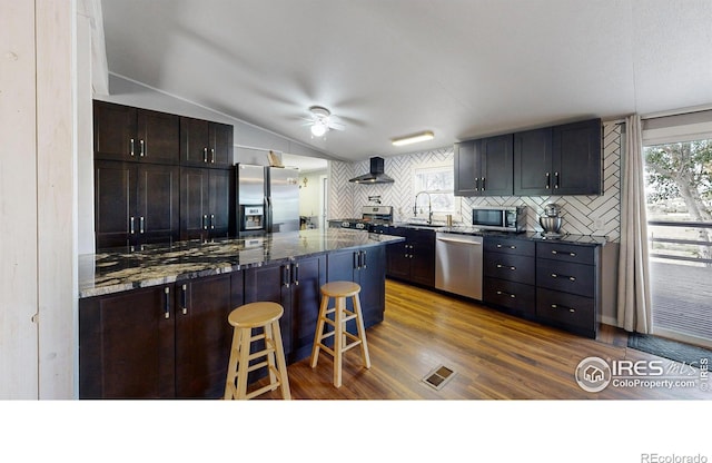 kitchen with stainless steel appliances, a sink, a kitchen breakfast bar, vaulted ceiling, and wall chimney exhaust hood