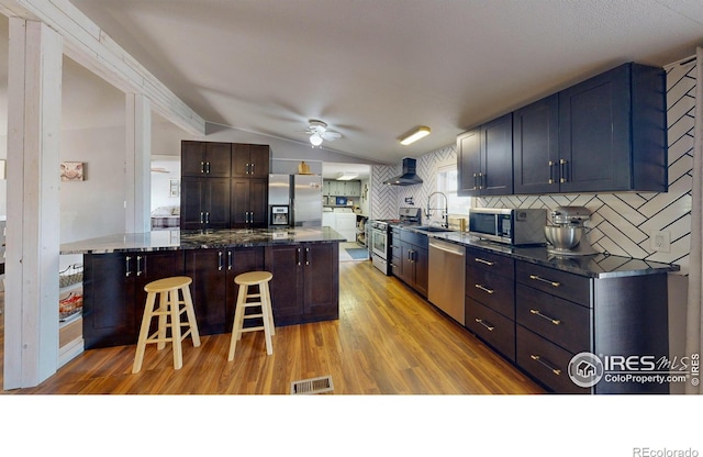 kitchen featuring a breakfast bar area, stainless steel appliances, vaulted ceiling, a sink, and wall chimney range hood