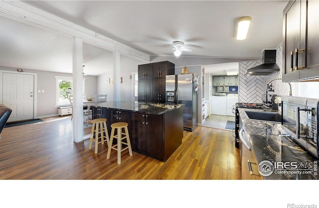 kitchen with washer and clothes dryer, a breakfast bar area, appliances with stainless steel finishes, vaulted ceiling, and wall chimney range hood