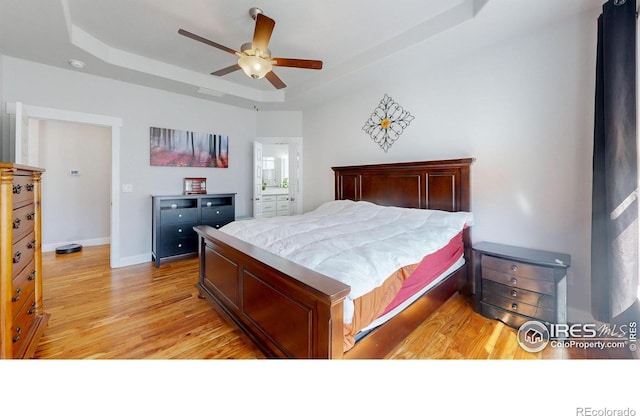 bedroom featuring a tray ceiling, light wood-style flooring, a ceiling fan, ensuite bath, and baseboards