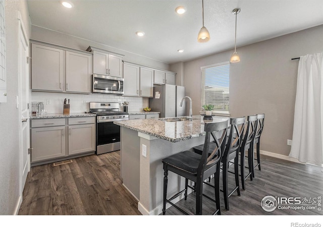 kitchen featuring an island with sink, light stone countertops, pendant lighting, stainless steel appliances, and dark hardwood / wood-style flooring