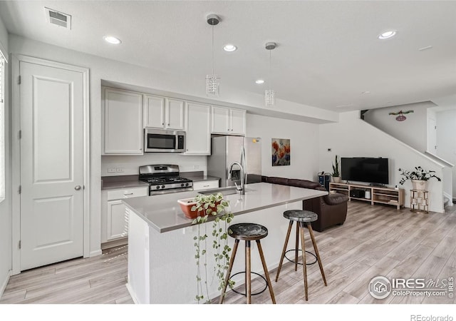 kitchen with light wood-type flooring, an island with sink, white cabinets, hanging light fixtures, and stainless steel appliances