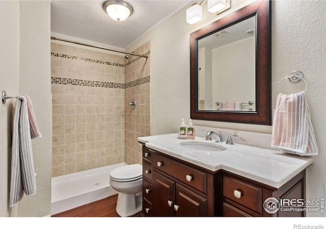 bathroom featuring a tile shower, hardwood / wood-style flooring, crown molding, vanity, and toilet