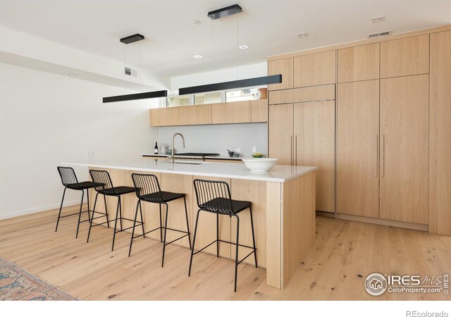 kitchen featuring light wood-type flooring, hanging light fixtures, a kitchen island with sink, and light brown cabinets