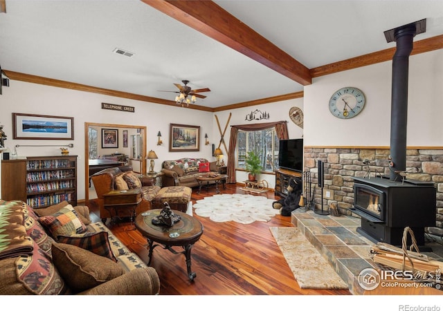 living room with visible vents, ceiling fan, wood finished floors, a wood stove, and beam ceiling