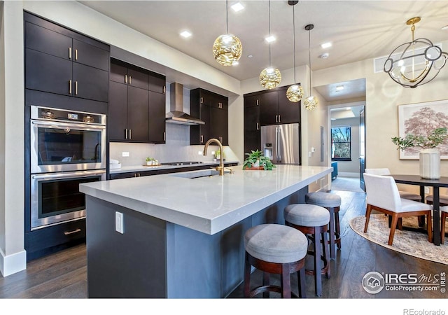 kitchen featuring pendant lighting, wall chimney range hood, appliances with stainless steel finishes, and a kitchen island with sink