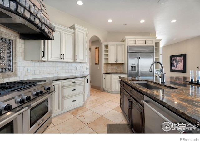 kitchen with ventilation hood, high quality appliances, dark stone countertops, sink, and decorative backsplash