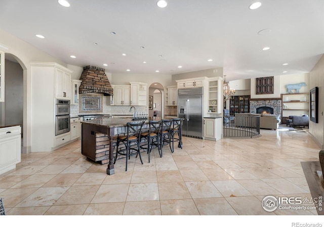 kitchen with tasteful backsplash, custom range hood, dark stone countertops, a large island with sink, and appliances with stainless steel finishes