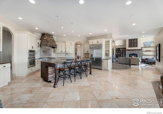 kitchen with stainless steel appliances, premium range hood, a stone fireplace, a breakfast bar area, and a large island with sink