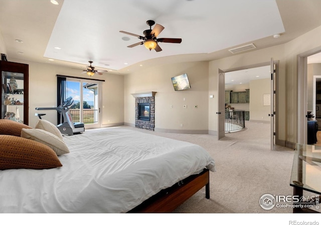 bedroom with a tray ceiling, ceiling fan, a stone fireplace, and light carpet