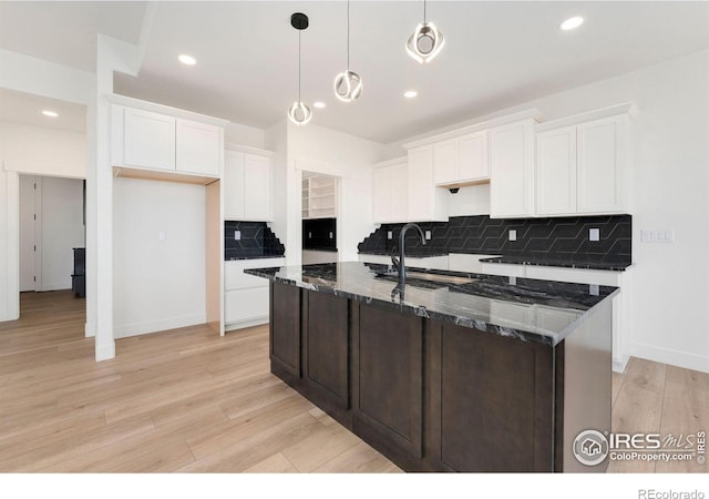 kitchen featuring dark stone counters, an island with sink, light wood-type flooring, white cabinets, and a sink