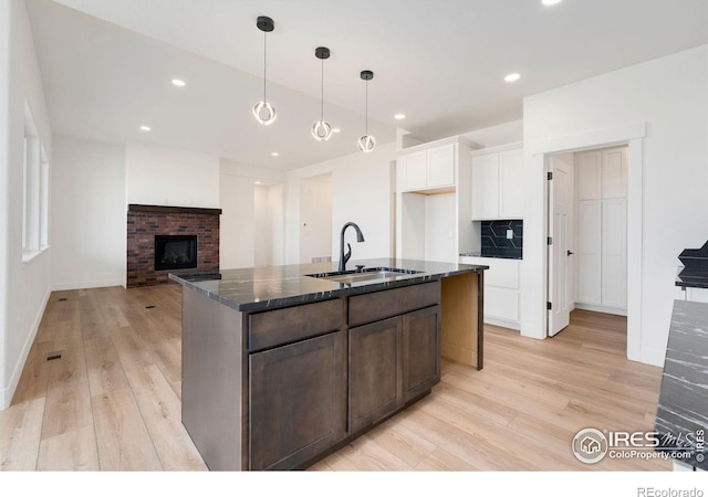 kitchen with a brick fireplace, light wood-type flooring, dark stone countertops, white cabinetry, and a sink