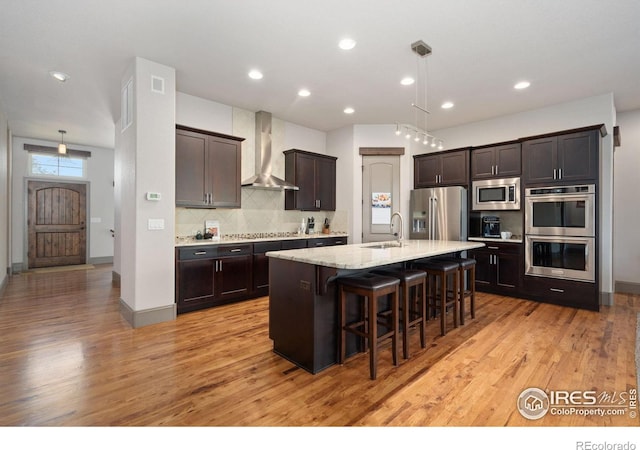 kitchen featuring an island with sink, wall chimney exhaust hood, appliances with stainless steel finishes, a breakfast bar, and light wood-type flooring