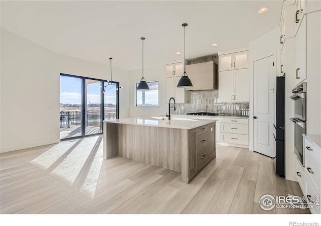 kitchen with an island with sink, backsplash, white cabinets, light hardwood / wood-style flooring, and sink