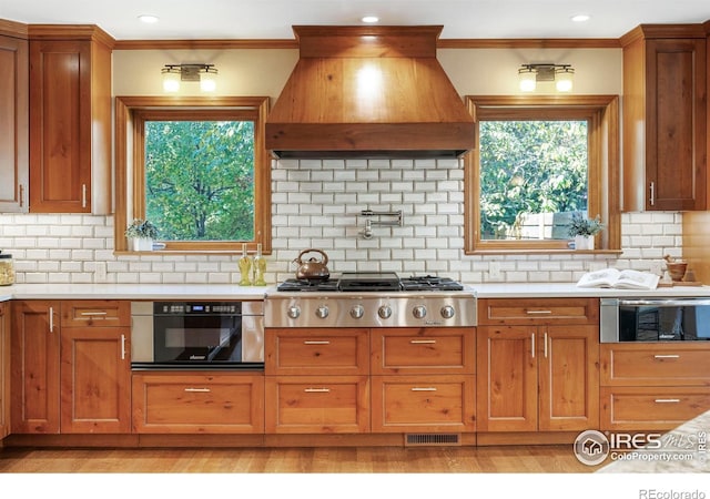 kitchen featuring custom range hood, backsplash, ornamental molding, appliances with stainless steel finishes, and light hardwood / wood-style floors