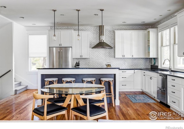 kitchen with wall chimney range hood, stainless steel appliances, hanging light fixtures, and a wealth of natural light