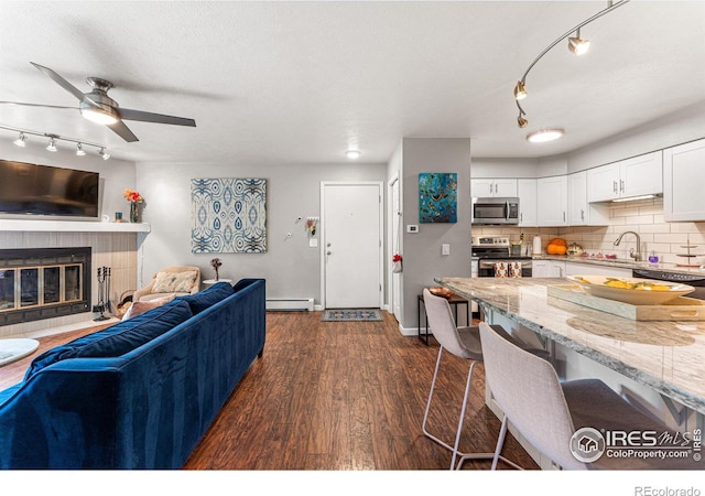 living room featuring a baseboard radiator, sink, a tiled fireplace, ceiling fan, and dark wood-type flooring