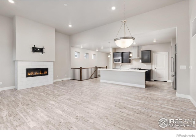 kitchen featuring wall chimney exhaust hood, an island with sink, stainless steel appliances, decorative light fixtures, and light hardwood / wood-style floors