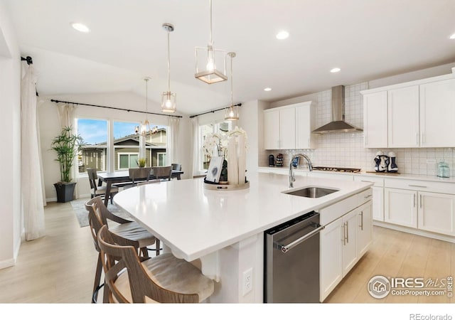 kitchen with wall chimney range hood, white cabinets, an island with sink, dishwasher, and decorative light fixtures