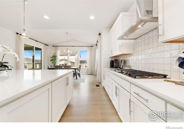 kitchen with wall chimney range hood, stainless steel gas cooktop, hanging light fixtures, lofted ceiling, and white cabinets