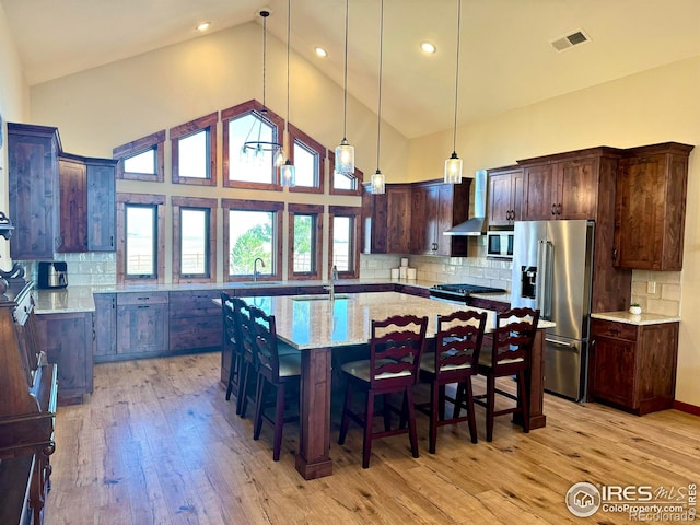 kitchen with decorative backsplash, appliances with stainless steel finishes, a kitchen island with sink, light stone countertops, and light wood-type flooring