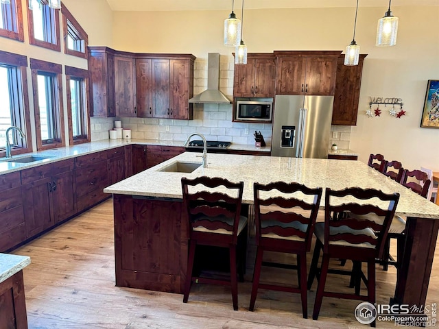 kitchen featuring appliances with stainless steel finishes, sink, wall chimney exhaust hood, light hardwood / wood-style flooring, and a kitchen island with sink