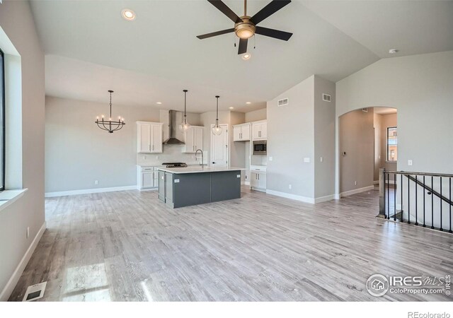 kitchen with wall chimney range hood, a kitchen island with sink, white cabinetry, light hardwood / wood-style floors, and decorative light fixtures