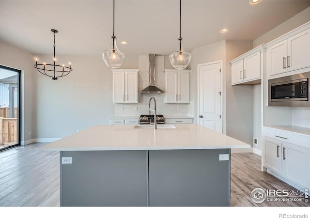 kitchen with stainless steel microwave, decorative light fixtures, white cabinetry, an island with sink, and wall chimney range hood